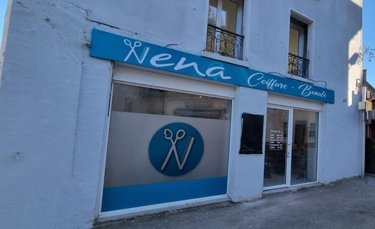 Une devanture blanche avec une enseigne bleue indiquant Nena Coiffure - Beauté au-dessus d'une vitrine ornée d'un logo circulaire représentant des ciseaux.