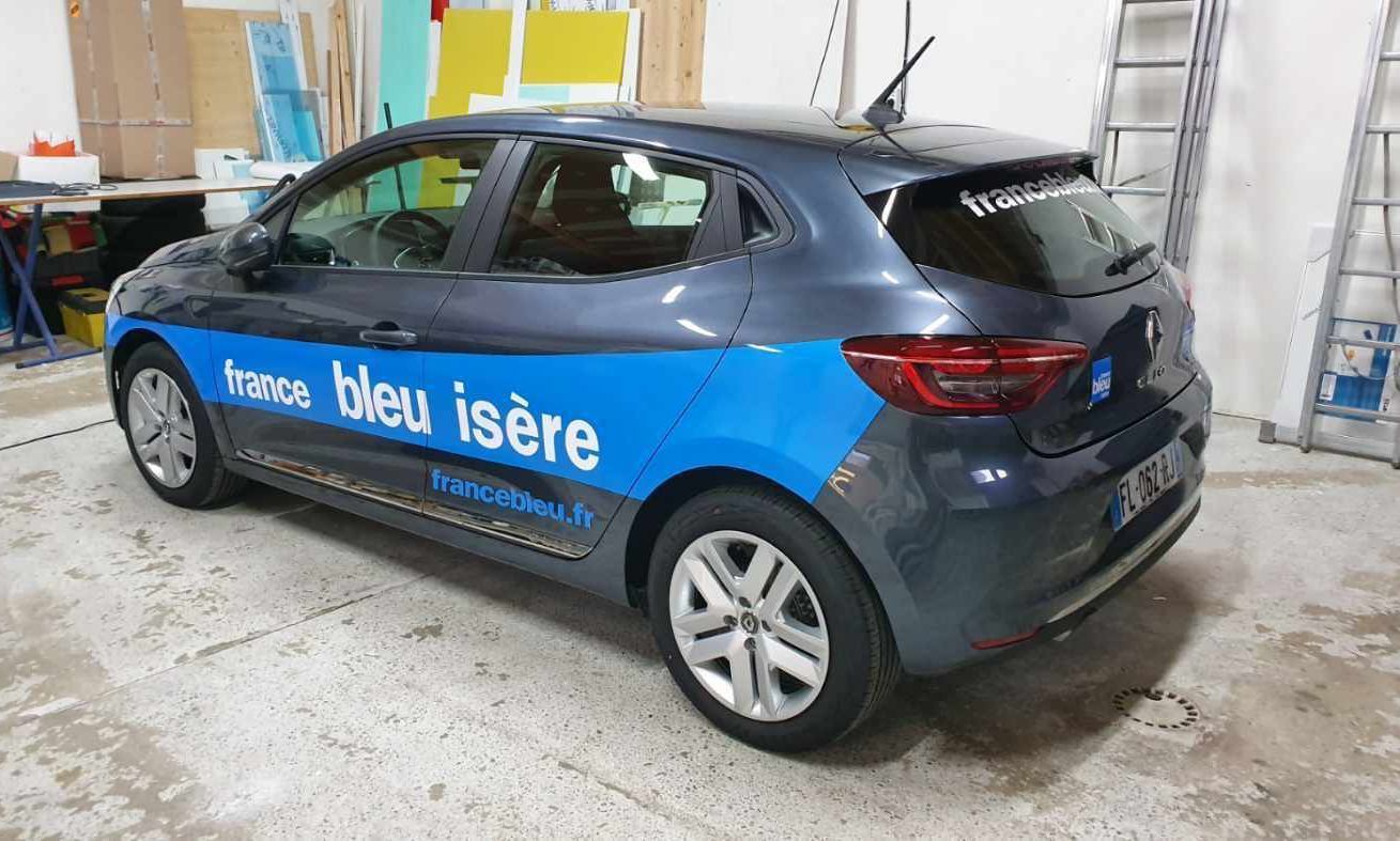 Voiture bleue portant l'inscription France Bleu Isère garée à l'intérieur, près d'une échelle.