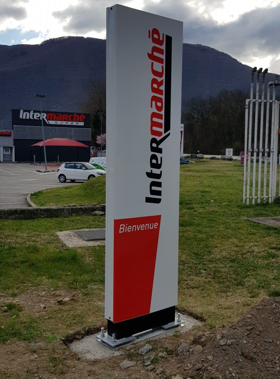 Enseigne Intermarché, haute et verticale, avec logo blanc et rouge, Bienvenue à la base, sur fond de montagne.