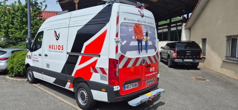 Une camionnette de marque Helios, ornée de graphismes géométriques blancs, noirs et rouges, est garée sur un parking extérieur à côté d'un pick-up noir.