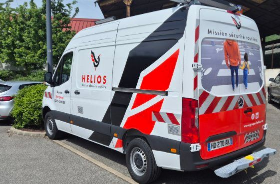 Une camionnette de livraison Helios blanche est garée à l'extérieur, ornée de motifs géométriques rouges et noirs et d'un pictogramme de sécurité routière à l'arrière.