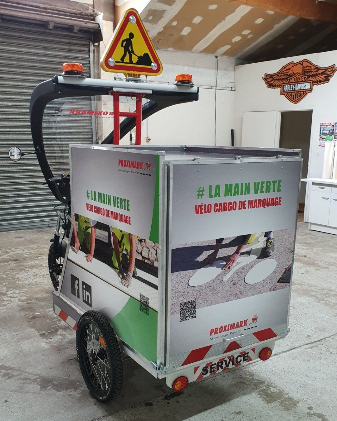 Un vélo cargo à trois roues avec un panneau de travaux et des feux clignotants.