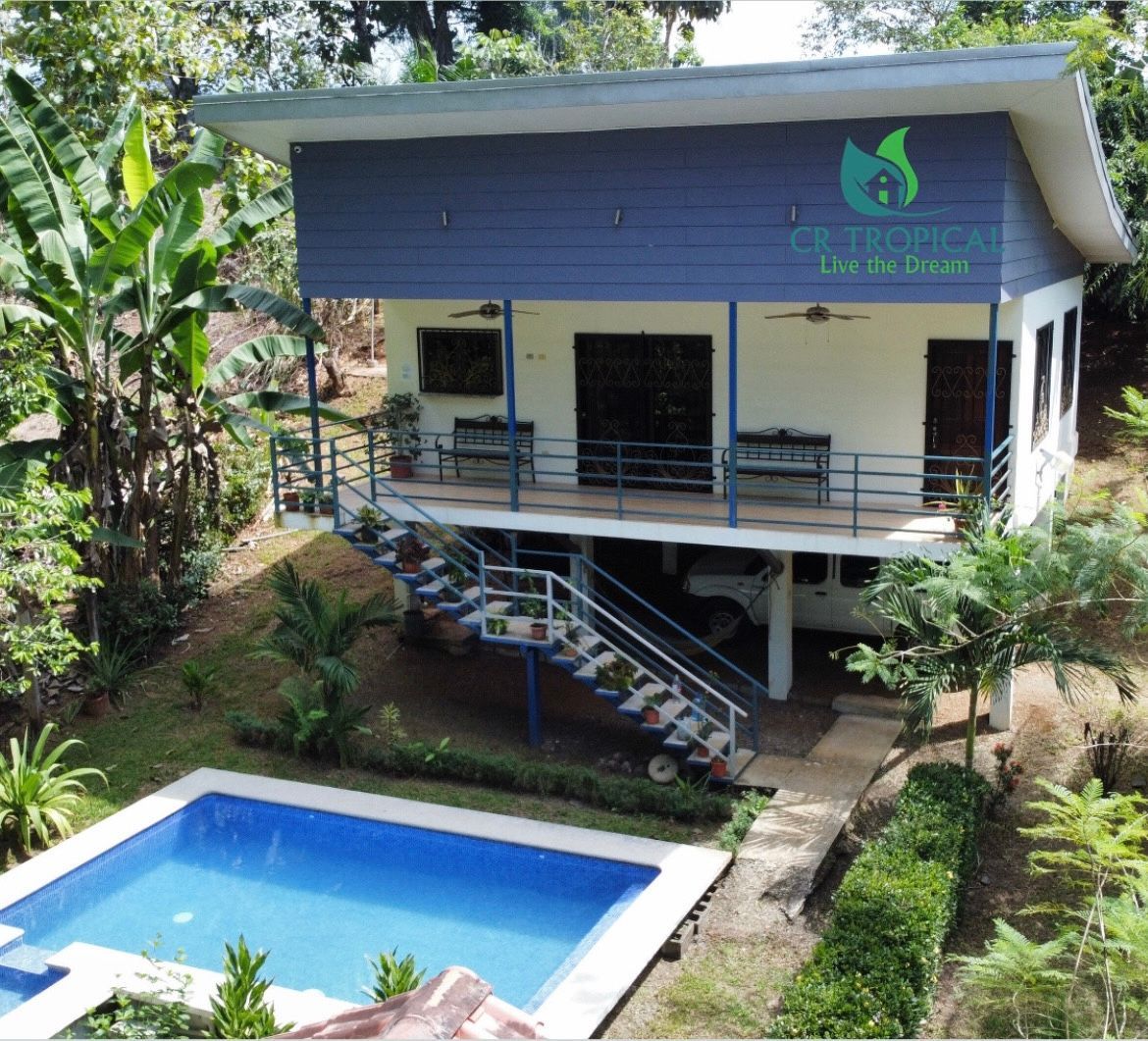 Two-story white house with blue roof, balcony, and a small swimming pool in a lush garden.