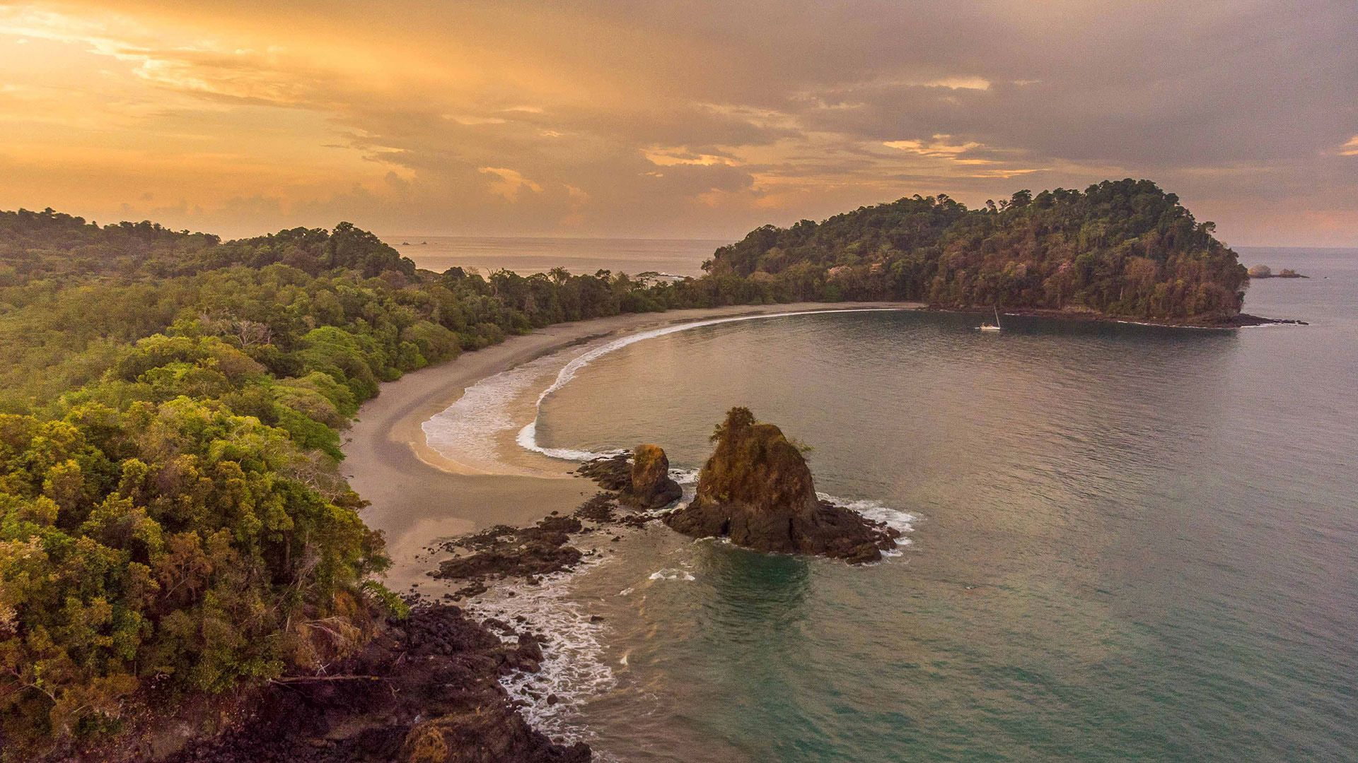 Sunset over a tropical bay with a crescent beach, rocky islet, and lush green coastline
