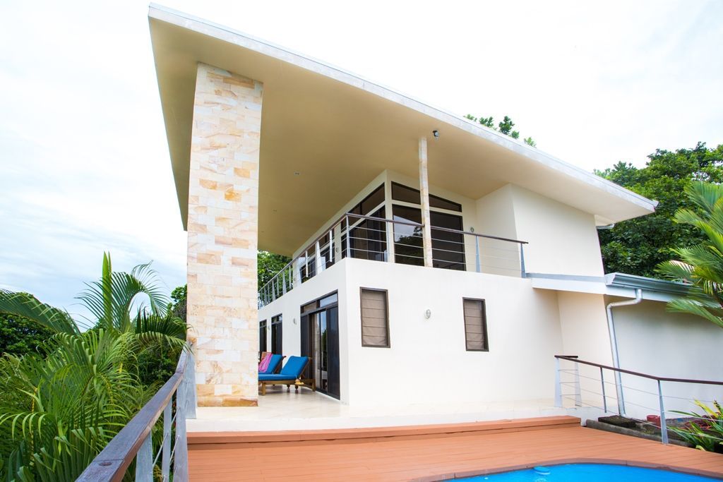 Modern white two-story house with stone column, large windows, wooden deck, and tropical greenery