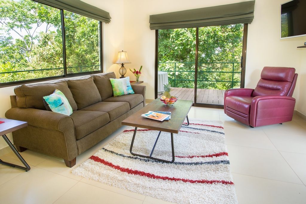 Bright living room with brown sofa, pink chair, coffee table, and large windows overlooking greenery.