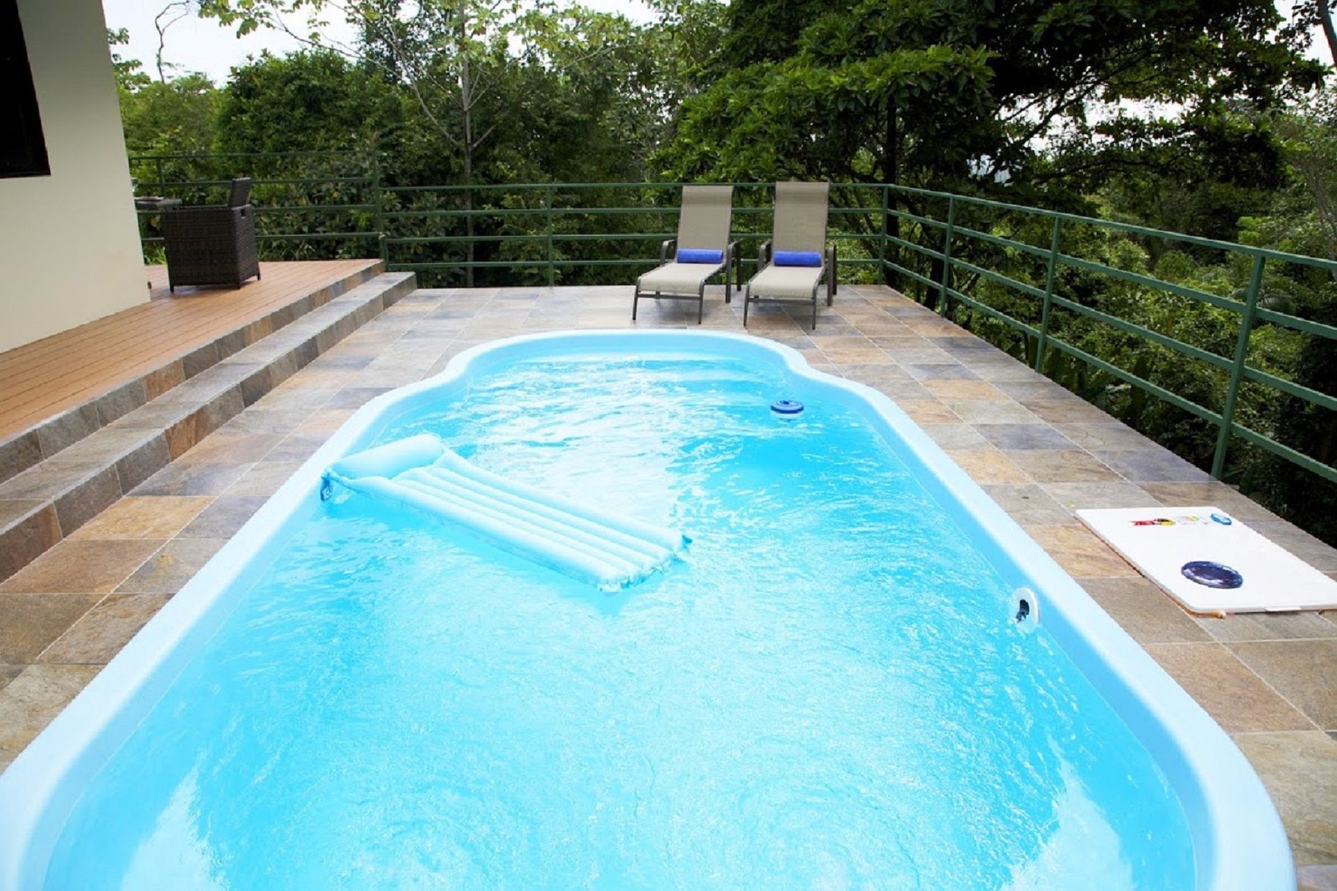 Small backyard pool with steps, lounge chairs, and lush greenery around a tiled patio.