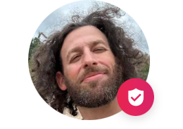 Close-up smiling man with curly hair and beard in a circular profile photo, with a pink badge icon in the corner