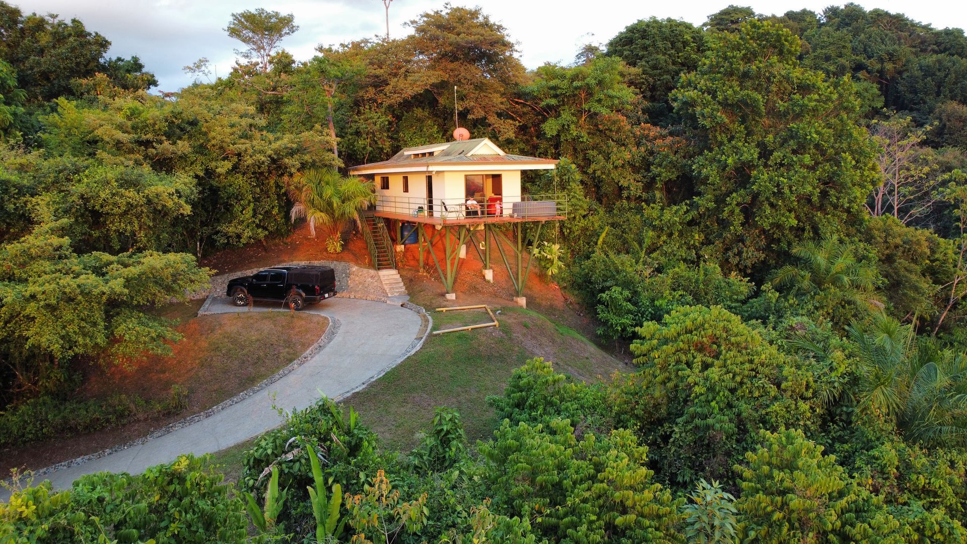Small house on a forested hillside with a winding driveway and a parked black car at sunset