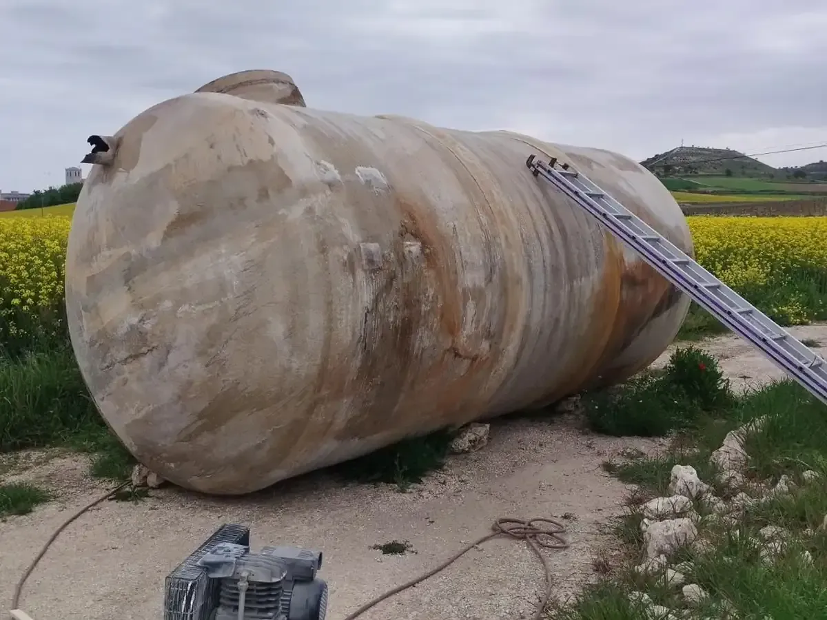 Gran tanque cilíndrico oxidado tirado al aire libre, con una escalera apoyada sobre él y un campo amarillo en el fondo.