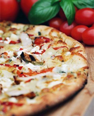 Una pizza está sobre una mesa de madera al lado de tomates y hojas de albahaca.