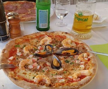 Una pizza con mariscos encima está sobre una mesa al lado de un vaso de cerveza.