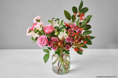 Strauß rosa und weißer Blumen mit grünem Laub in einem Glasgefäß auf einer weißen Oberfläche.