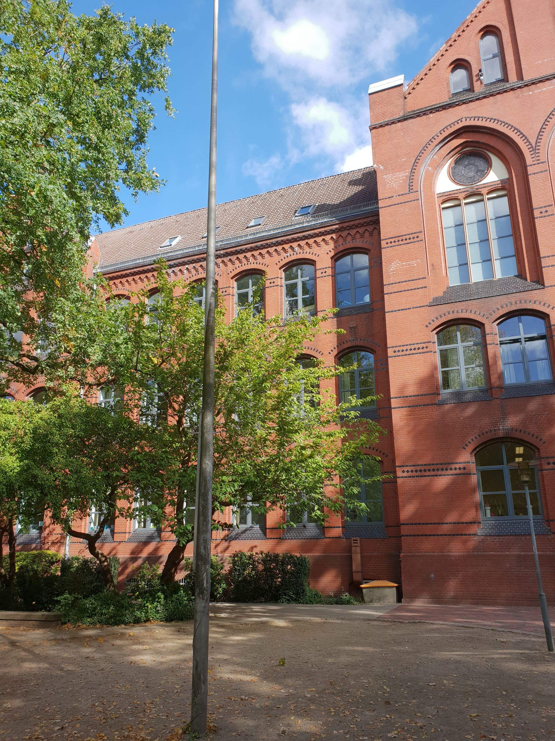 Rotes Backsteingebäude mit Bogenfenstern und einem Baum im Vordergrund an einem sonnigen Tag