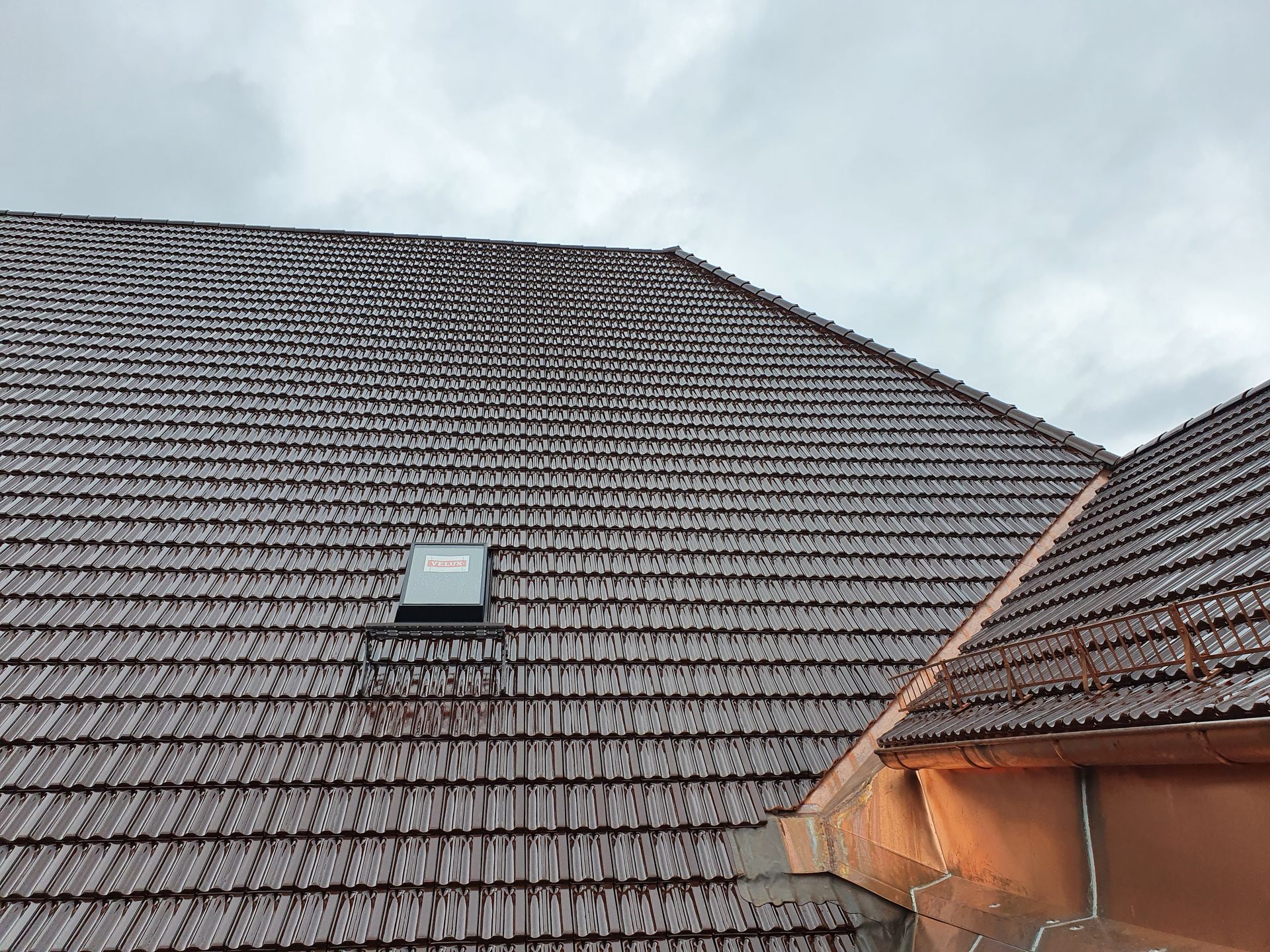 Dach mit braunen Ziegeln, ein Oberlicht und eine kleine überdachte Dachgaube unter einem bewölkten Himmel