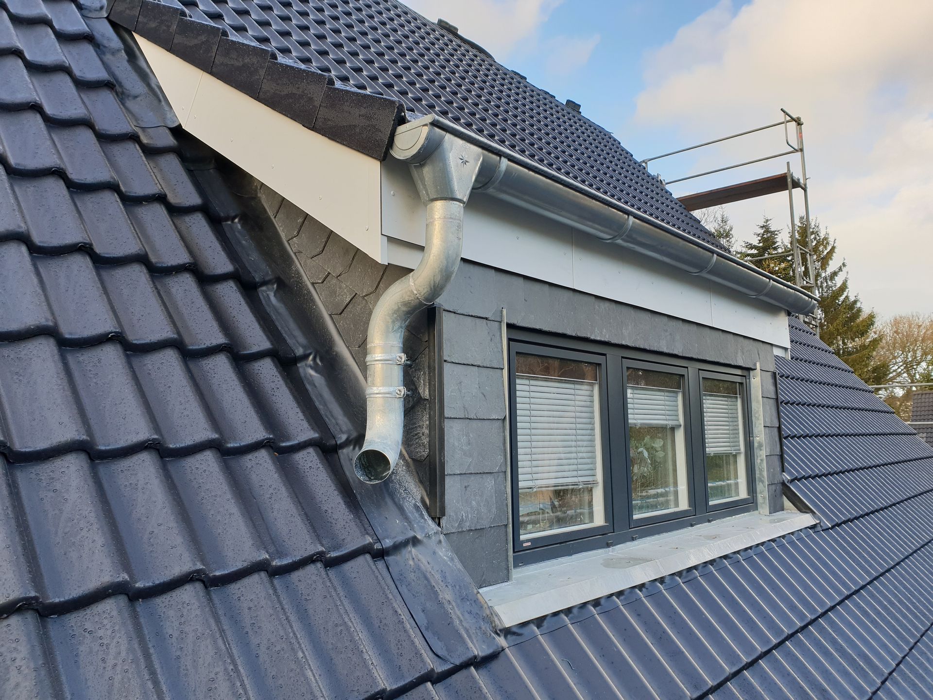 Dachgaubenfenster auf einem grauen Ziegeldach mit silbernem Fallrohr und Baugerüst im Hintergrund