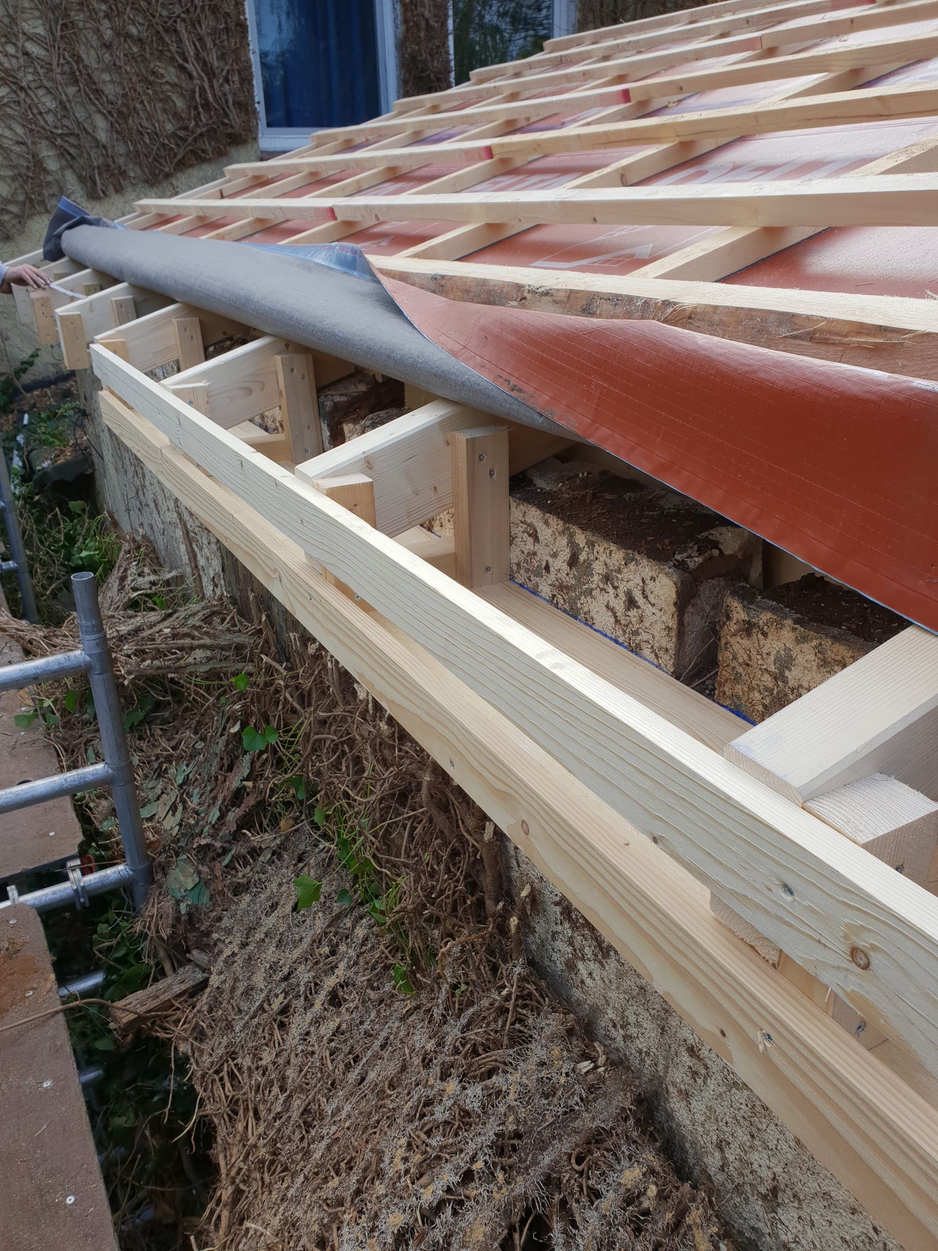 Teilweise errichtetes Dachgerüst mit Holzsparren und roten Dachplatten entlang einer Steinmauer