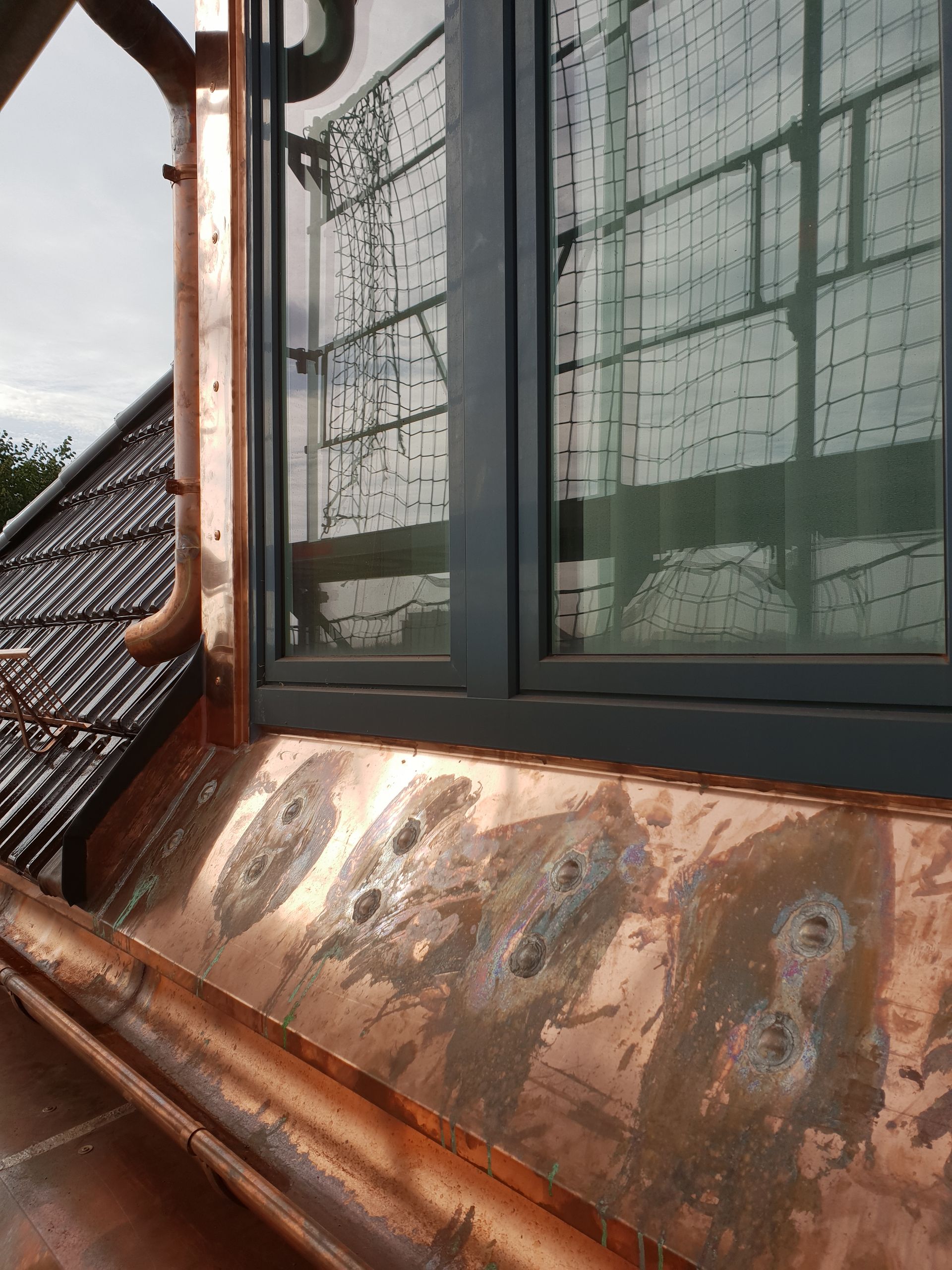 Rostiges Kupferdach unter einem Fenster mit grünen Vorhängen und einer Dachlinie im Hintergrund