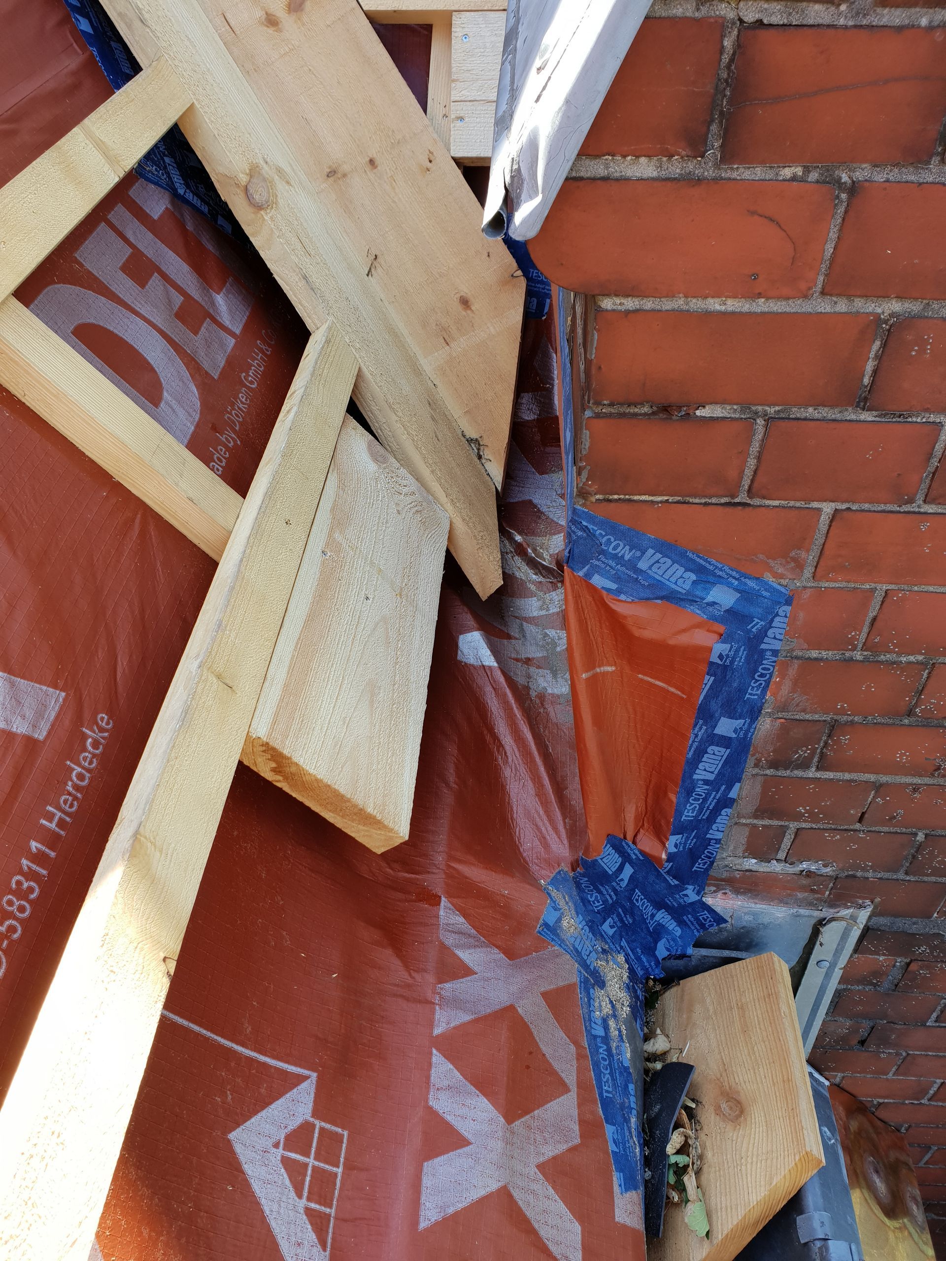 Zerbrochene Holzbretter und blaues Klebeband lehnen an einer Backsteinmauer neben roter Plastikfolie.
