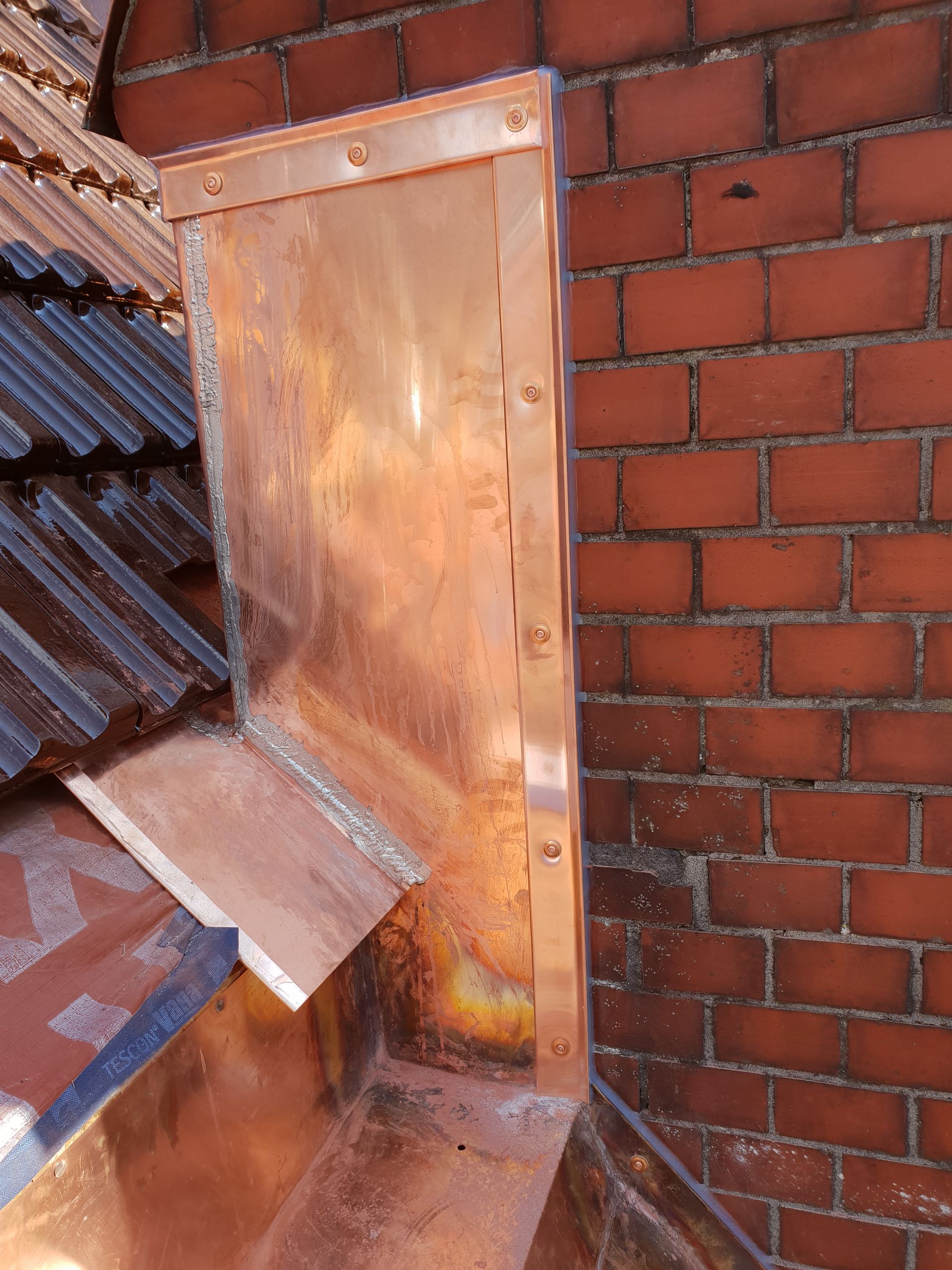 Metallene Kaminverkleidung und Entlüftung an einer roten Backstein-Dachwand neben dunklen Dachziegeln.