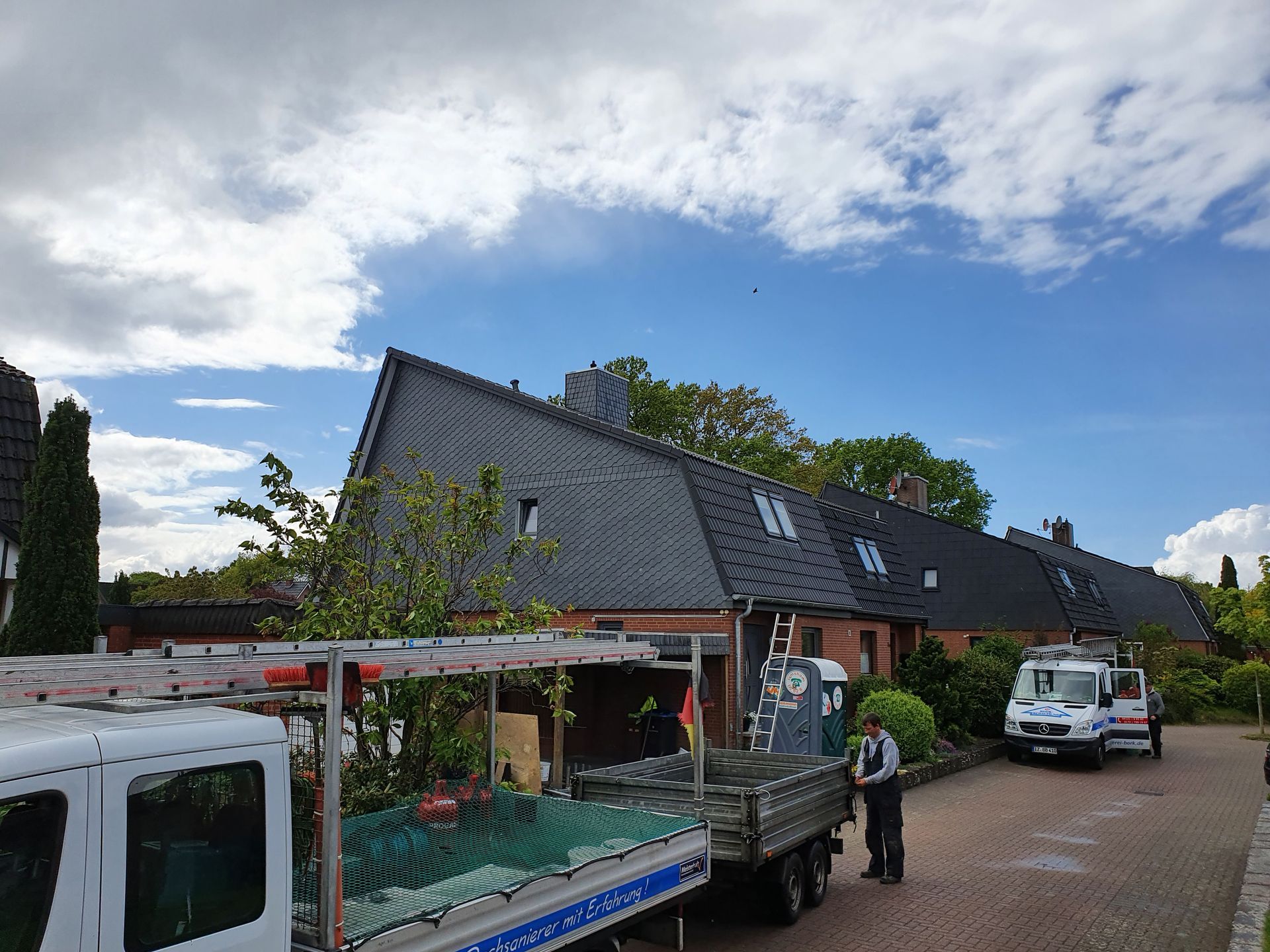 Backsteinhaus mit dunklem Dach, weiße Lieferwagen und ein Arbeiter, der unter einem bewölkten Himmel einen LKW in einer Einfahrt belädt