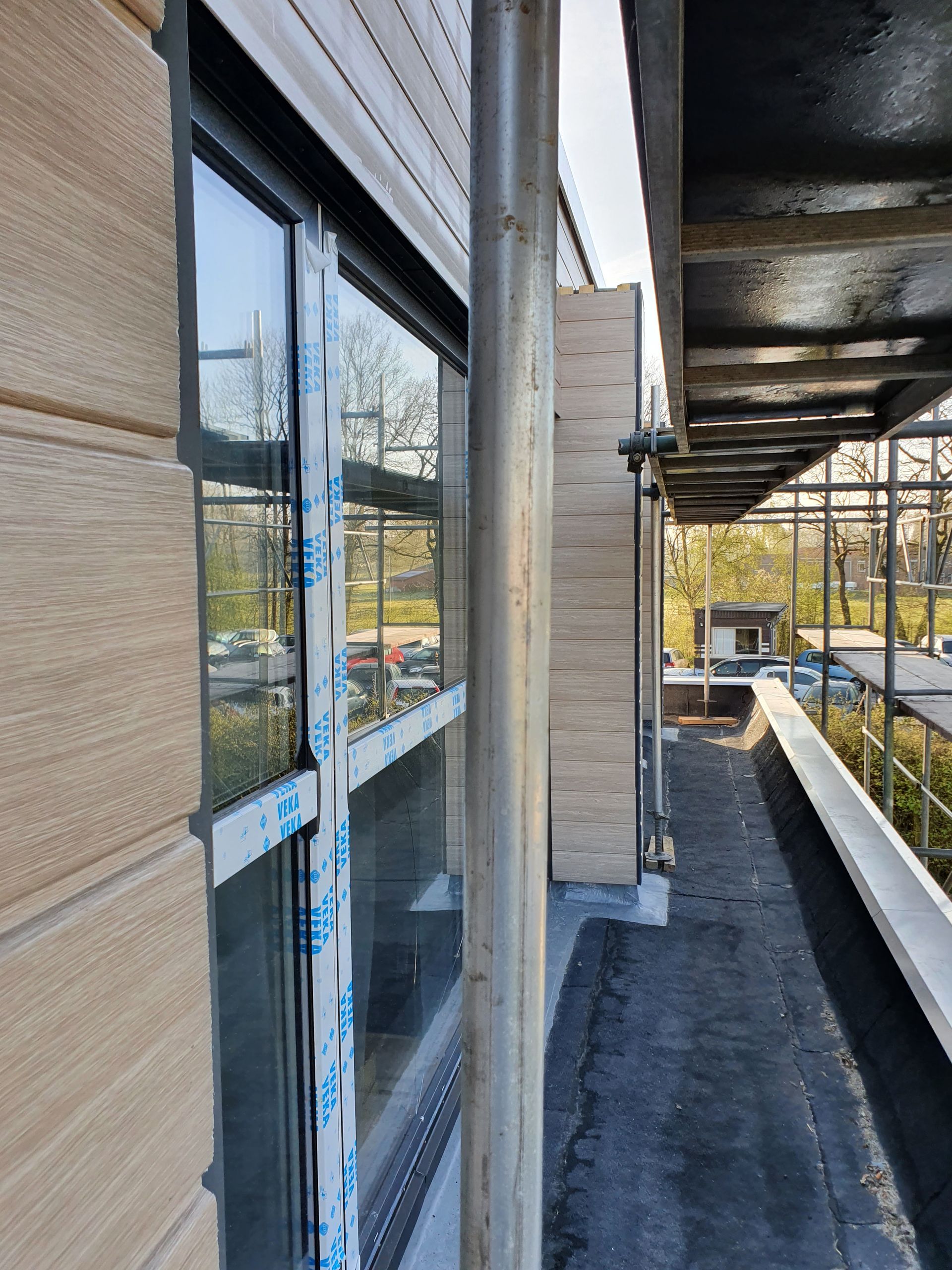 Schmaler Balkonsteg neben einem Gebäude, links Fenster, rechts ein Baugerüst.