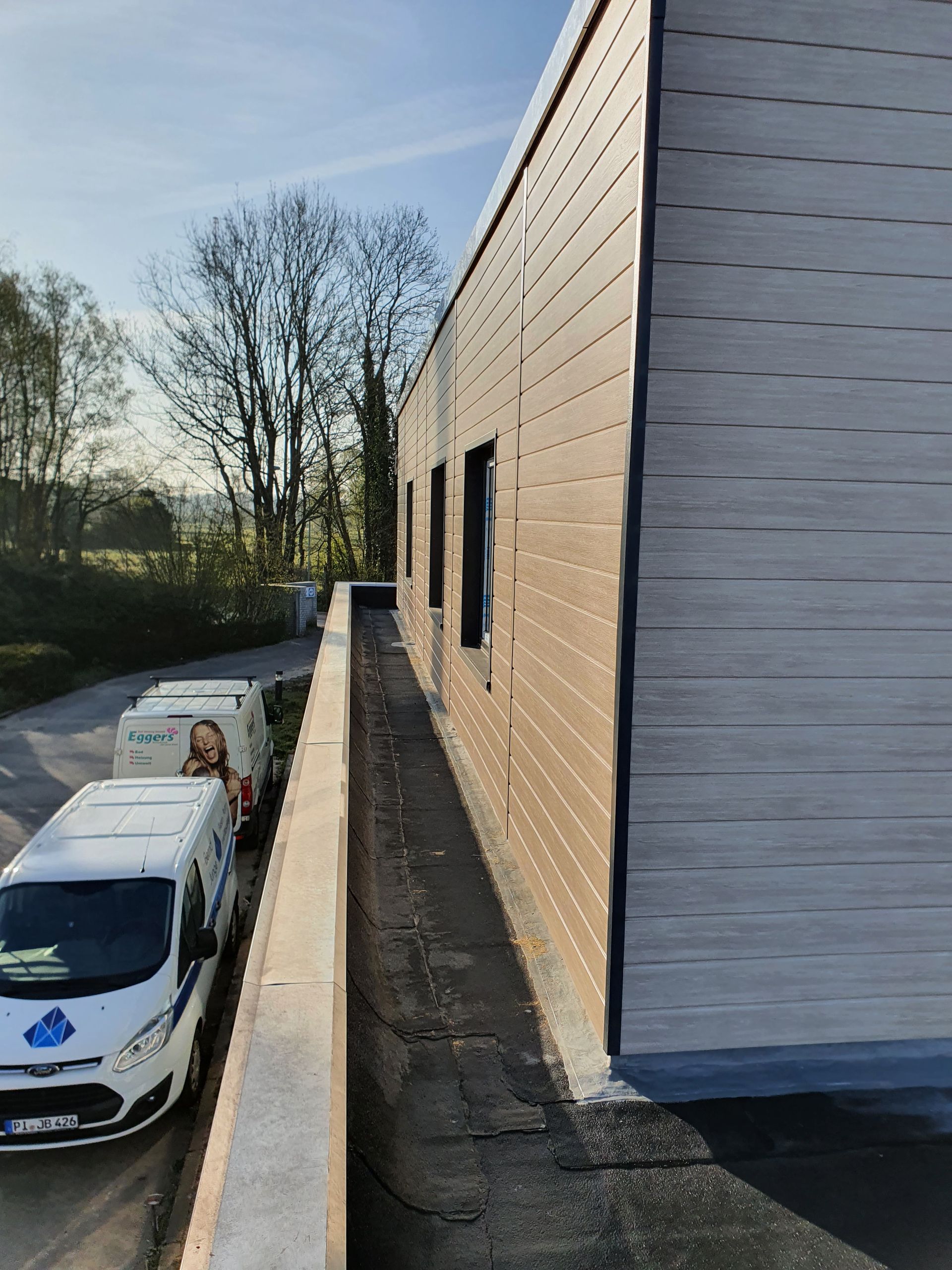 Ein Industriegebäude neben einem schmalen Weg, darunter geparkte Lieferwagen und in der Ferne Bäume.