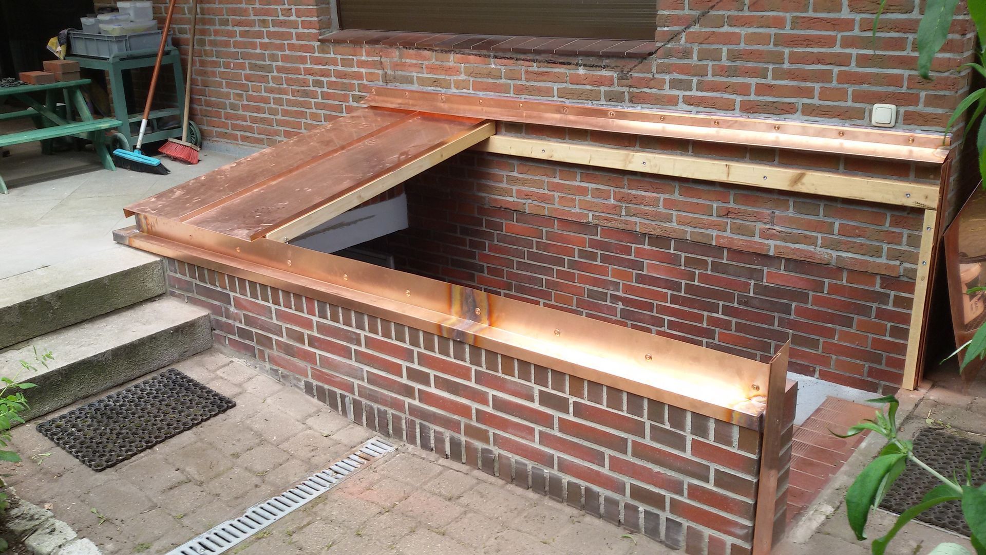 Eine mit Ziegeln ausgekleidete, tieferliegende Rampe oder ein Treppenhaus neben einem Gebäude, mit hölzerner Abdeckung und einem nahegelegenen Metallablaufgitter.
