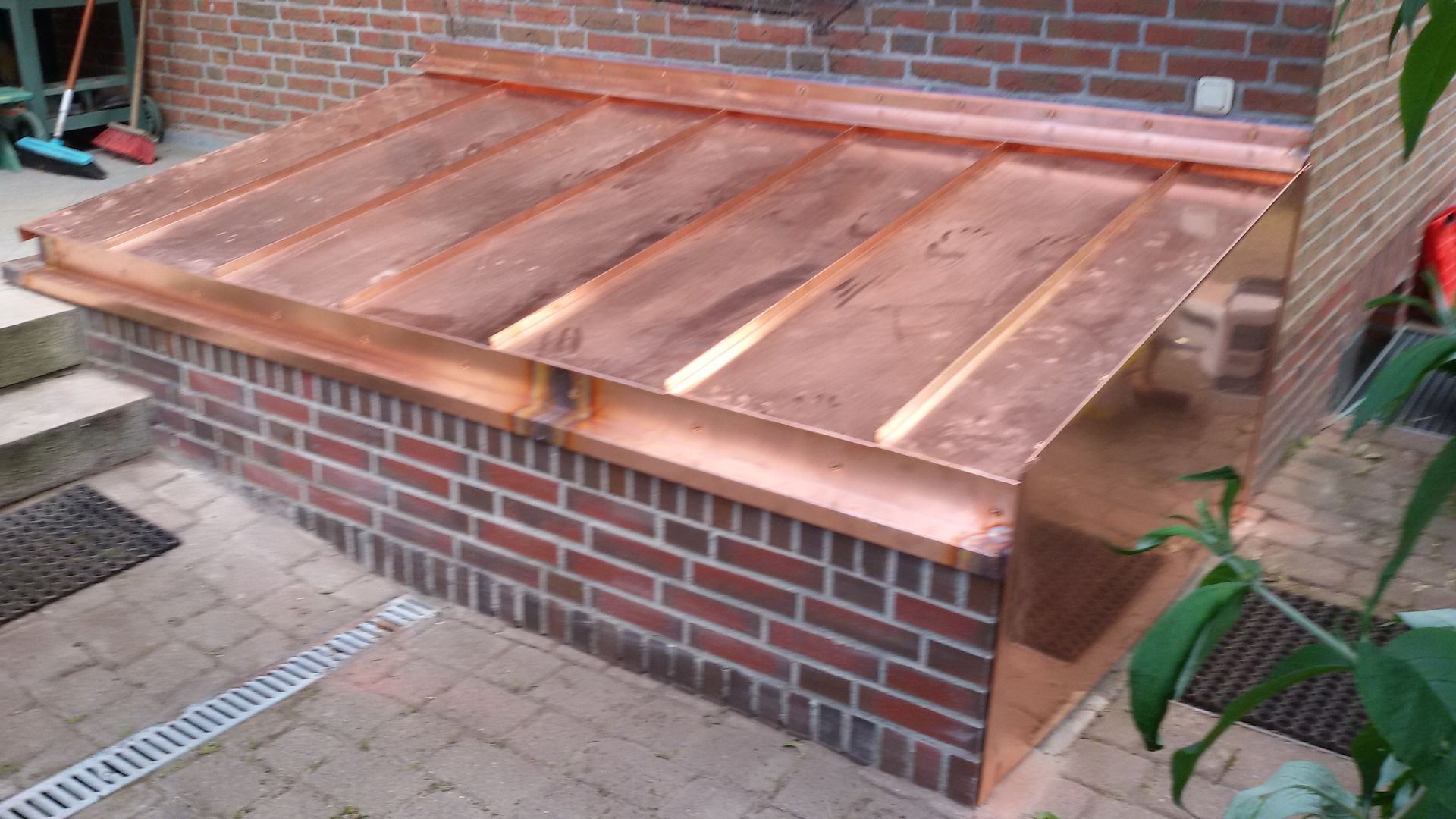 Ein gemauertes Außengehege mit schrägem Holzdeckel liegt neben einer Ziegelmauer und einer Terrasse.