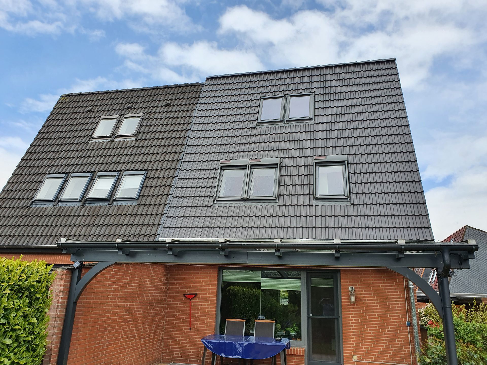 Modernes Backsteinhaus mit dunklem Ziegeldach, Oberlichtern und einer überdachten Terrasse mit blauen Stühlen