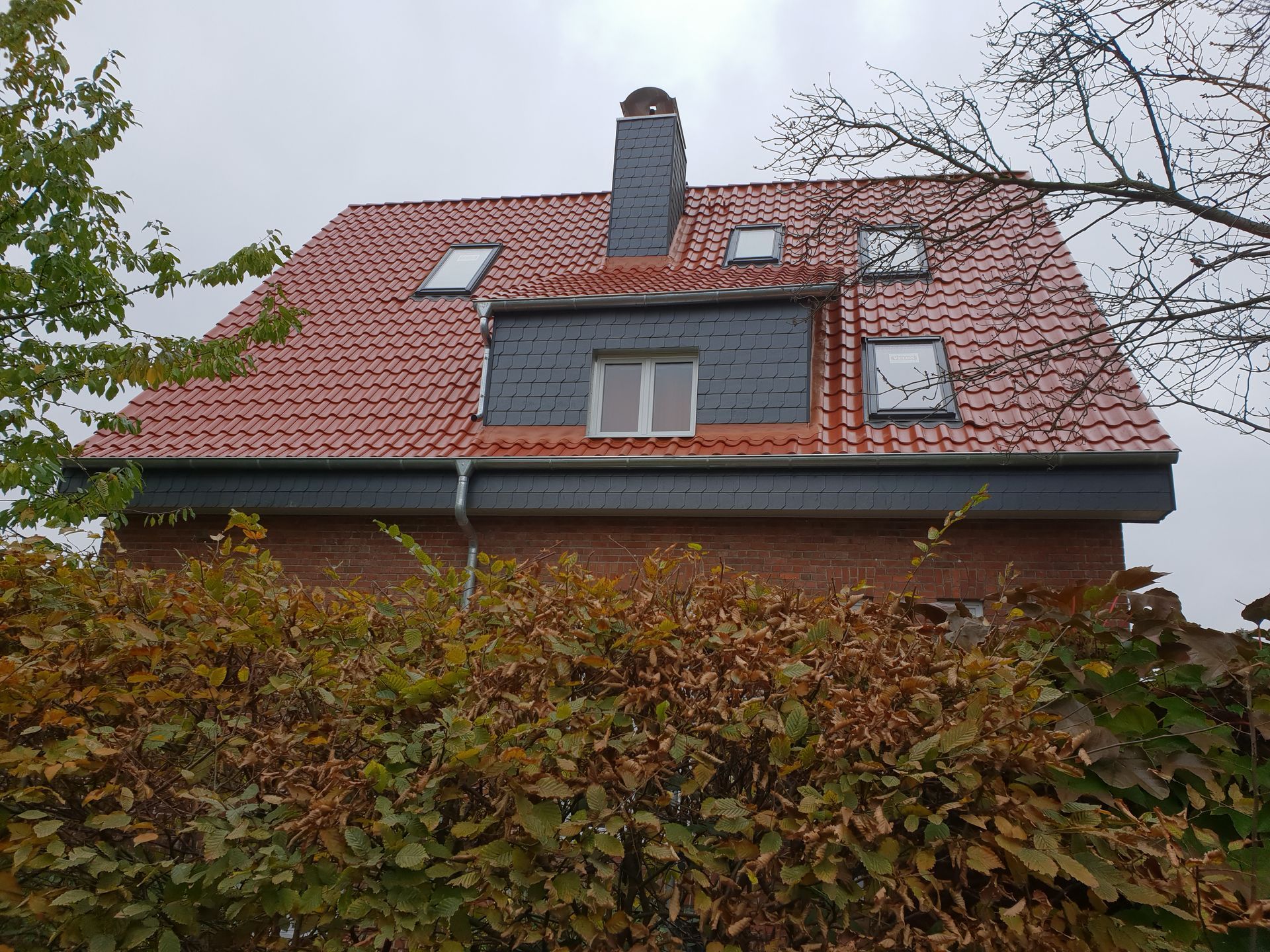 Haus mit rotem Dach, Dachgauben, Schornstein und efeubewachsener Mauer unter bedecktem Himmel