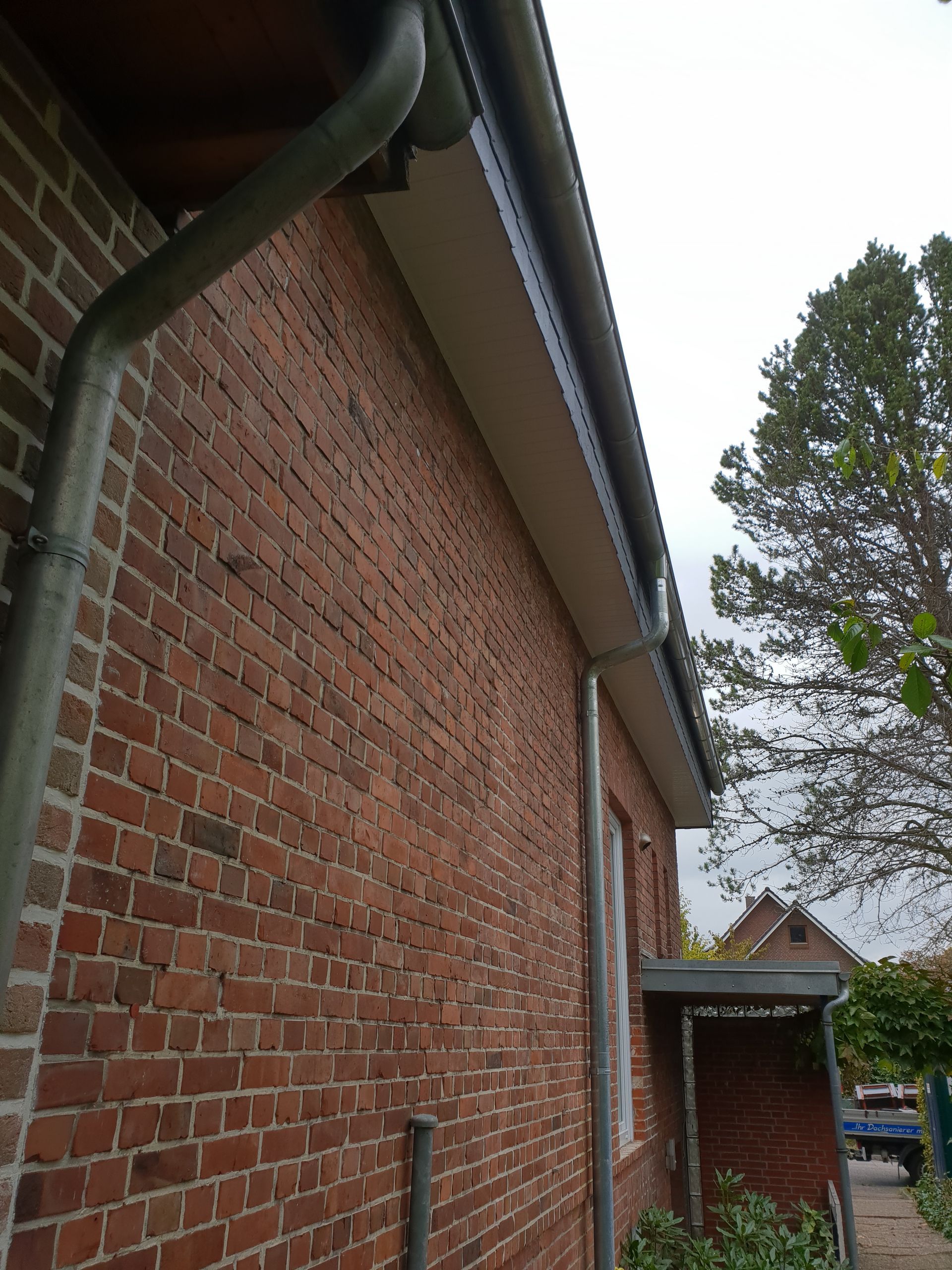 Eine Hauswand aus roten Ziegelsteinen mit Dachrinne und Dachrand neben einer kleinen Veranda unter bedecktem Himmel.