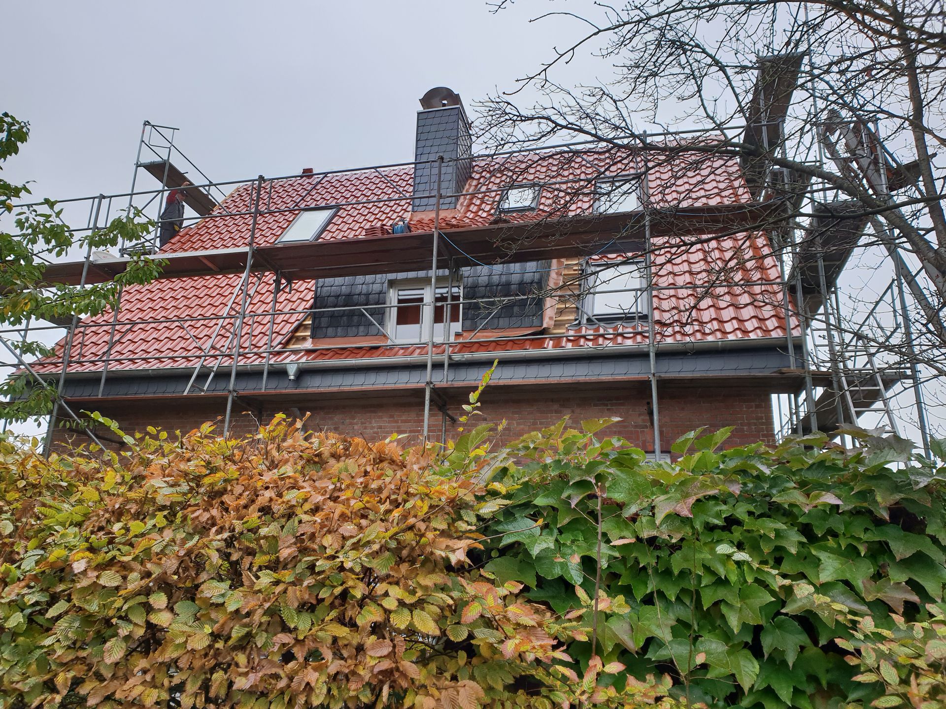 Teilweise überdachtes Haus mit rotem Dach und Gerüst, gesehen über grünem Herbstlaub