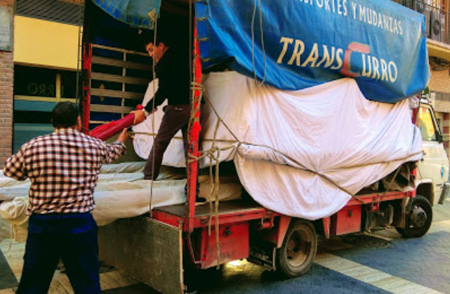 Dos hombres cargando un camión rojo con lonas blancas y letras azules, estacionado en una calle.