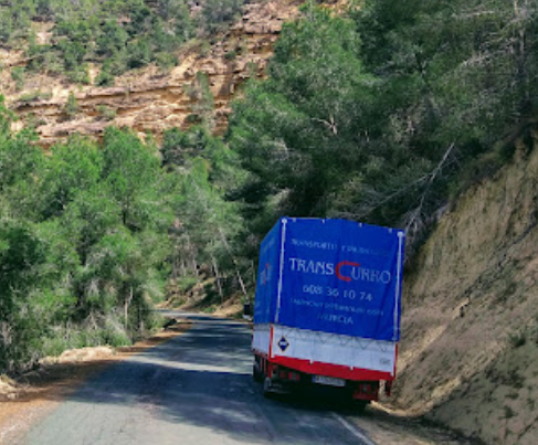 Camión en una carretera sinuosa en una zona montañosa, lona azul, rodeado de árboles y acantilados.