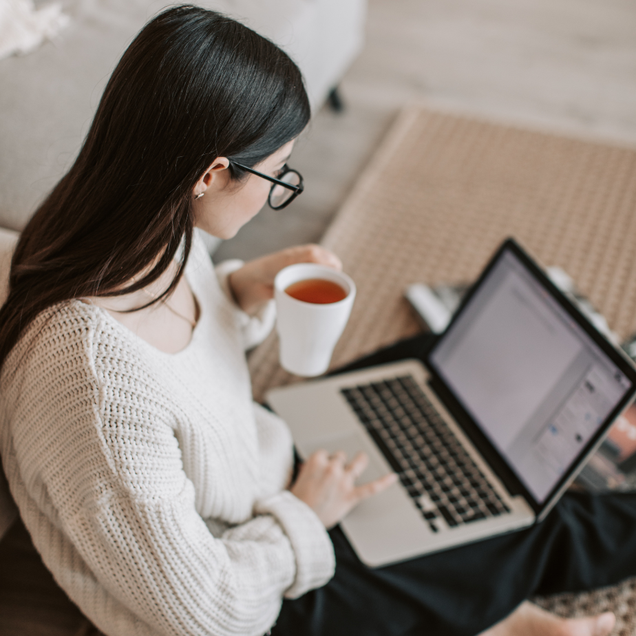 A woman is sitting on the floor using a laptop and drinking tea