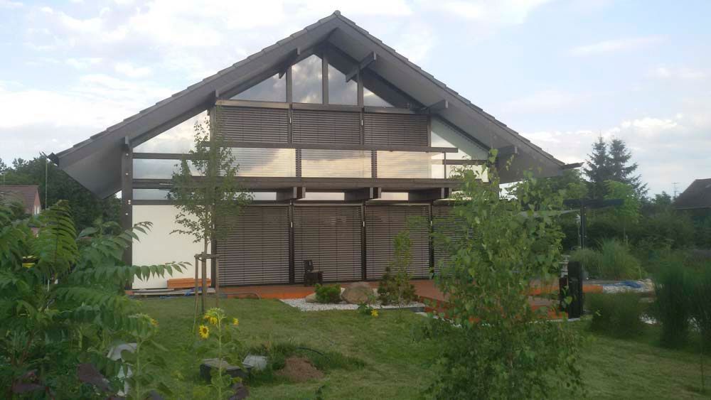 Modernes Haus mit Glaswänden und dunklem Schrägdach, umgeben von Grün und einem Garten.