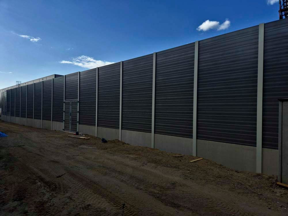 Eine lange, dunkle Schallschutzmauer neben einer unbefestigten Straße unter blauem Himmel.