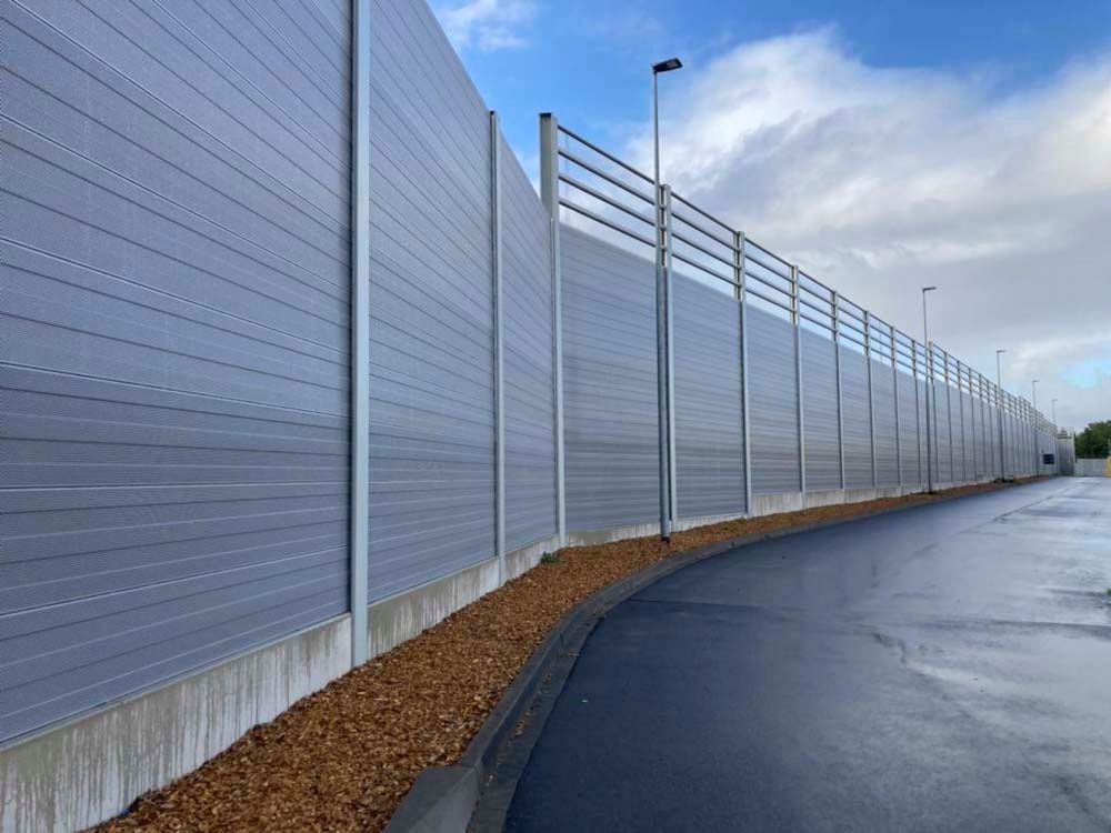 Eine lange, graue Lärmschutzwand entlang einer kurvigen Straße, darüber ein bewölkter Himmel.