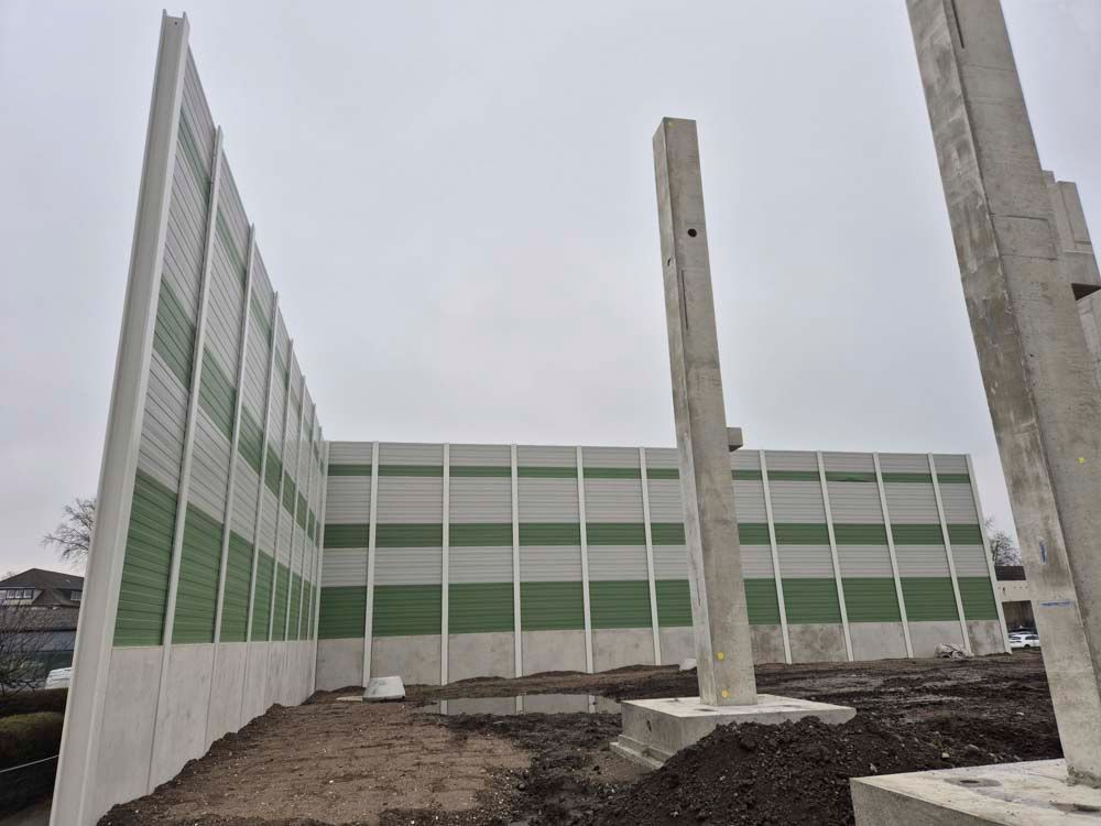 Baustelle mit Betonmauer und Säulen. Grüne und weiße Platten als Schallschutzwand. Bewölkter Tag.
