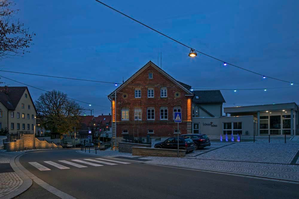 Ein Backsteingebäude in der Abenddämmerung mit Lichterketten darüber, in der Nähe eines Zebrastreifens und geparkter Autos.