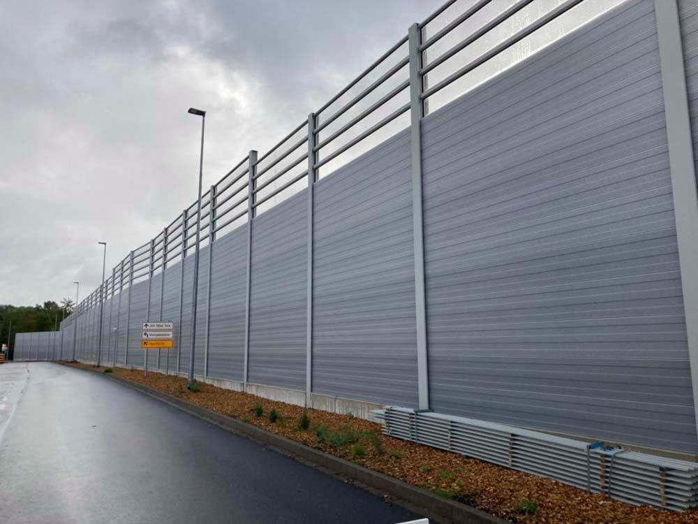 Lange graue Lärmschutzwand entlang einer Straße unter bewölktem Himmel.