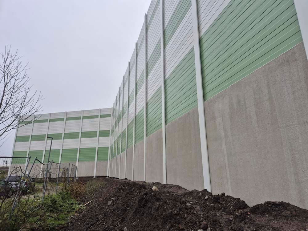 Hohe Lärmschutzwand mit abwechselnd grünen und weißen Paneelen vor einem grauen Himmel.