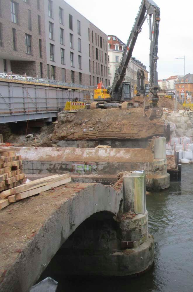 Baustelle: Bagger reißt Brücke über einen Wasserweg ab. Braune Erde, Steinpfeiler und Gebäude im Hintergrund.