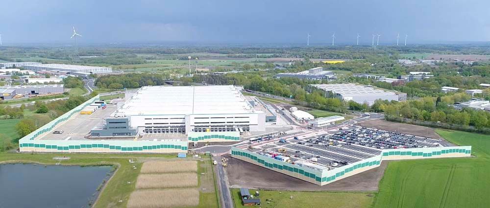 Luftaufnahme eines großen Lagerhauses mit einem grün-weißen Umzäunungszaun, umgeben von Feldern und einem Gewässer.