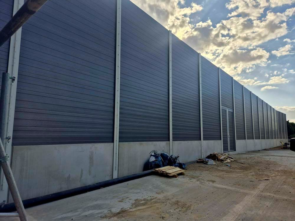 Eine lange, graue Lärmschutzwand auf einem Betonsockel mit einer Tür vor einem bewölkten Himmel.