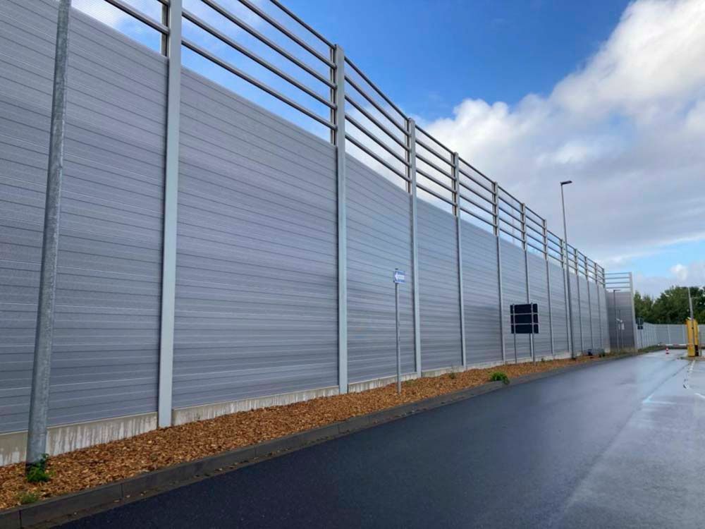 Graue Lärmschutzwand entlang einer Straße mit Metallpfosten und offenem oberen Abschnitt an einem bewölkten Tag.
