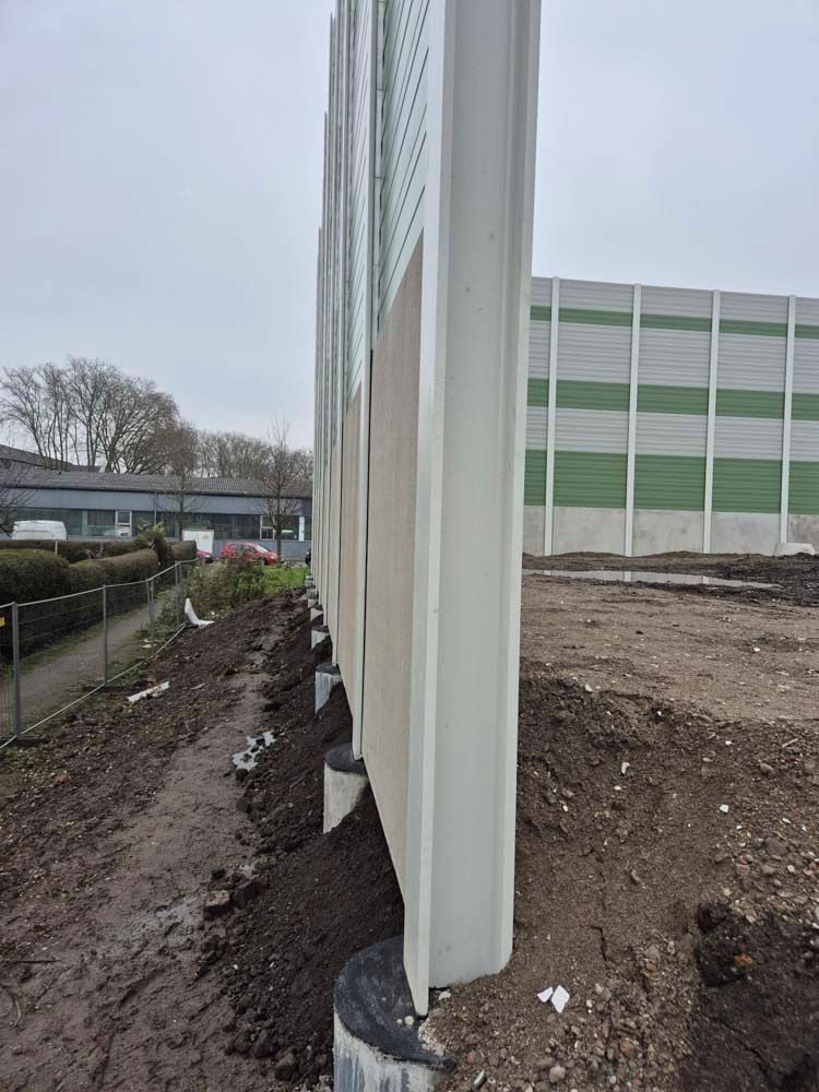 Lärmschutzwand entlang einer Straße mit weißen, grauen und grünen Paneelen. An der Basis befinden sich Erde und Pfosten.