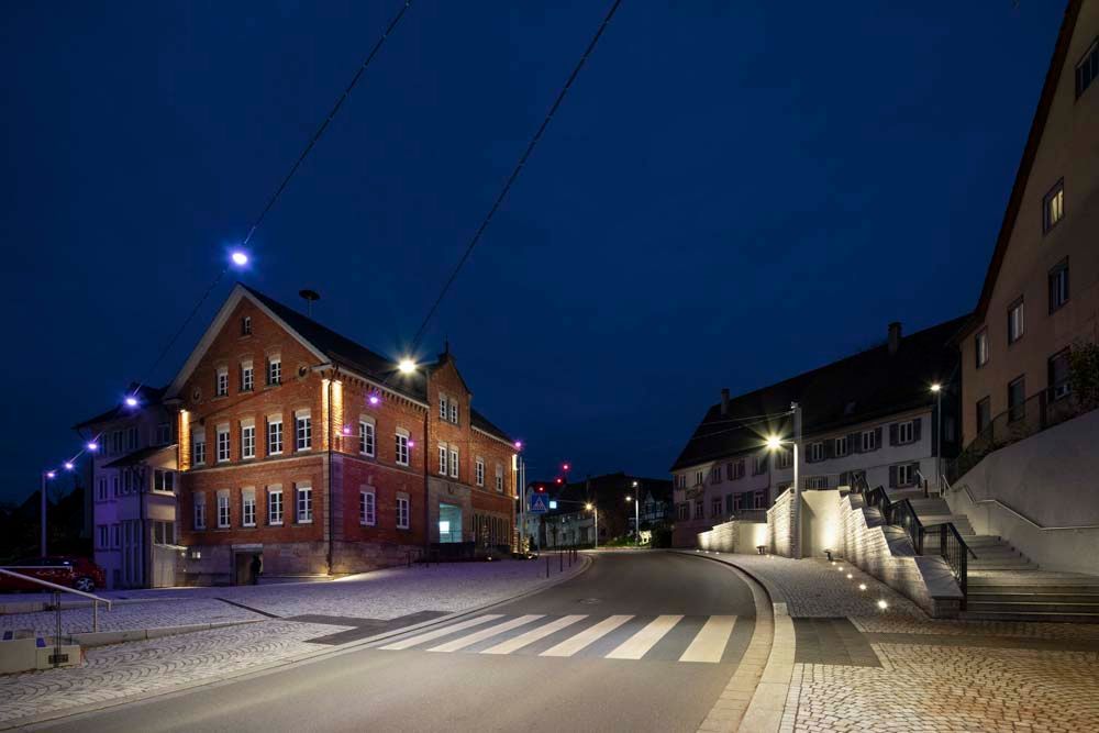 Nachtansicht einer Kopfsteinpflasterstraße in einer Stadt. Gebäude werden von Straßenlaternen beleuchtet, Zebrastreifen sichtbar. Dunkelblauer Himmel.