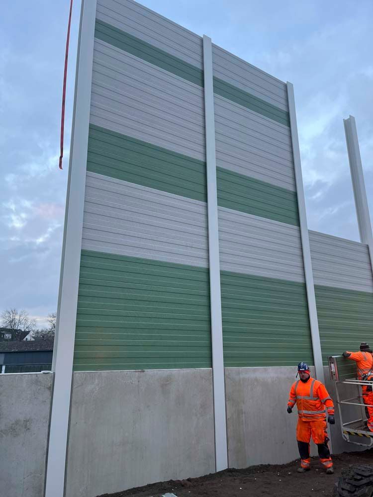 Bauarbeiter installieren eine hohe, grün-graue Lärmschutzwand neben einem Betonfundament.